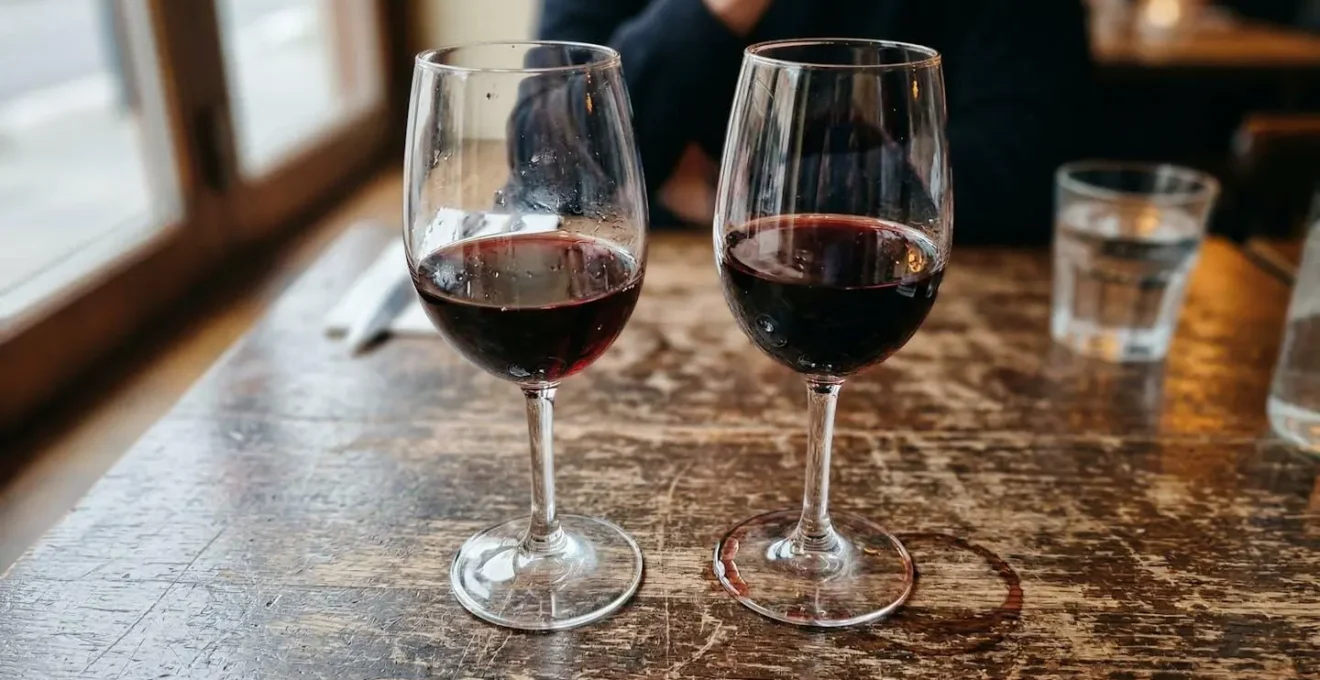Deux verres à vin posés côte à côte sur une table en bois rustique, l'un visiblement plus fin que l'autre sous la lumière latérale